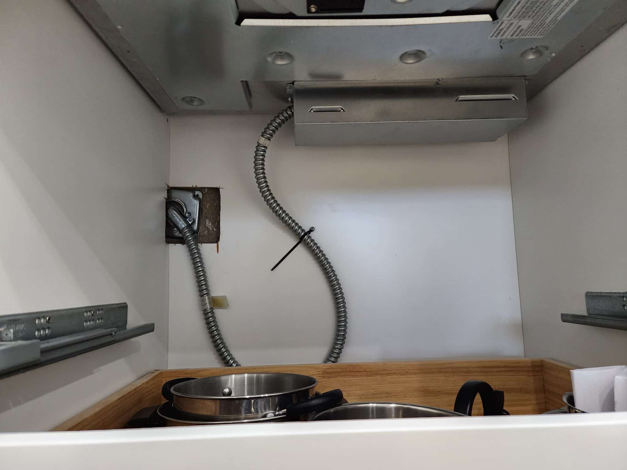 Interior view of a lower kitchen cabinet showing the armored electrical cable and wall junction box that supplies high-voltage power to the built-in electric cooking appliance.