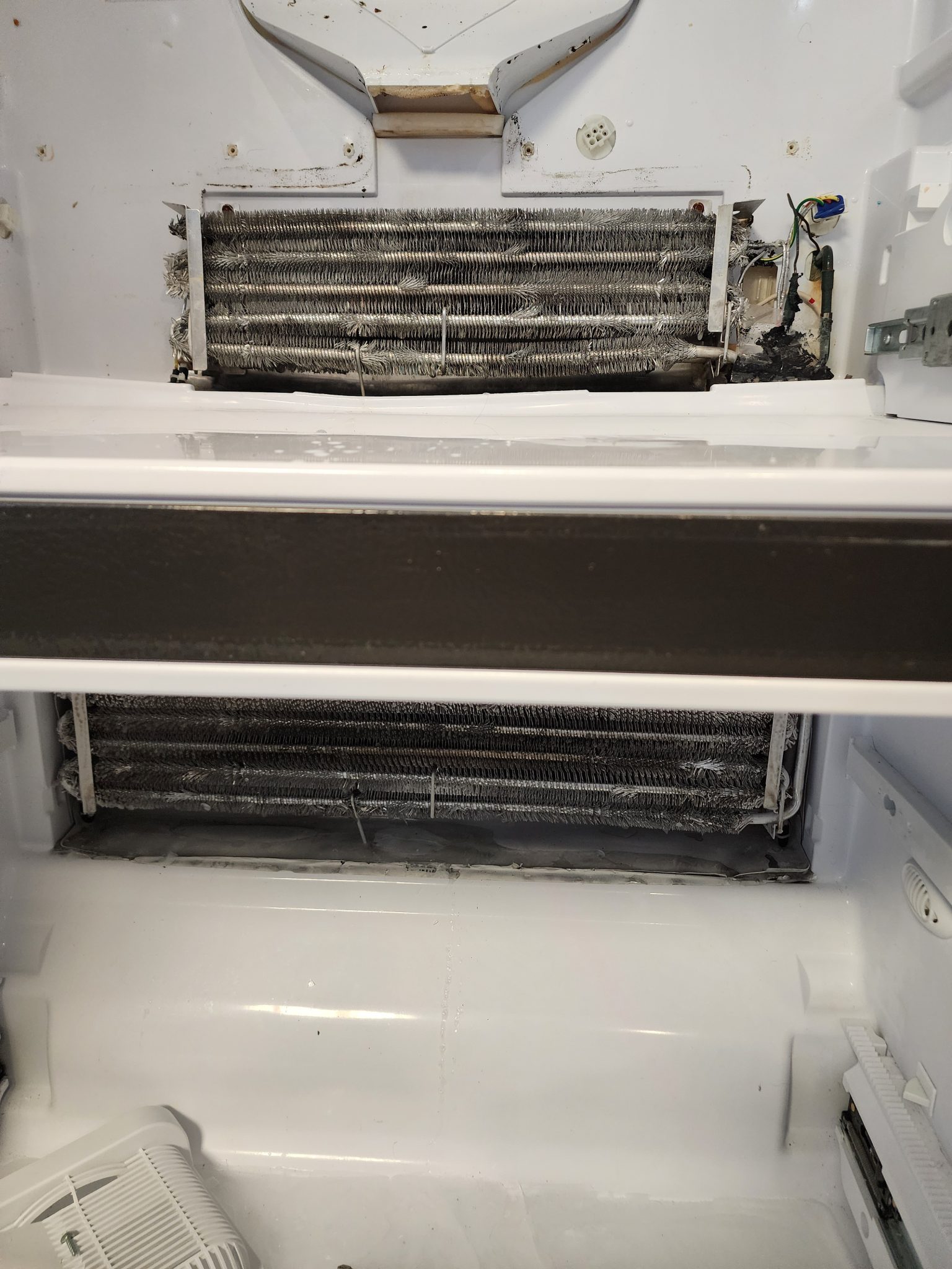 Interior view of a disassembled GE Profile refrigerator showing two stacked radiator-style evaporator coils with severe electrical burn damage and melted wiring on the far right side of the upper coil.